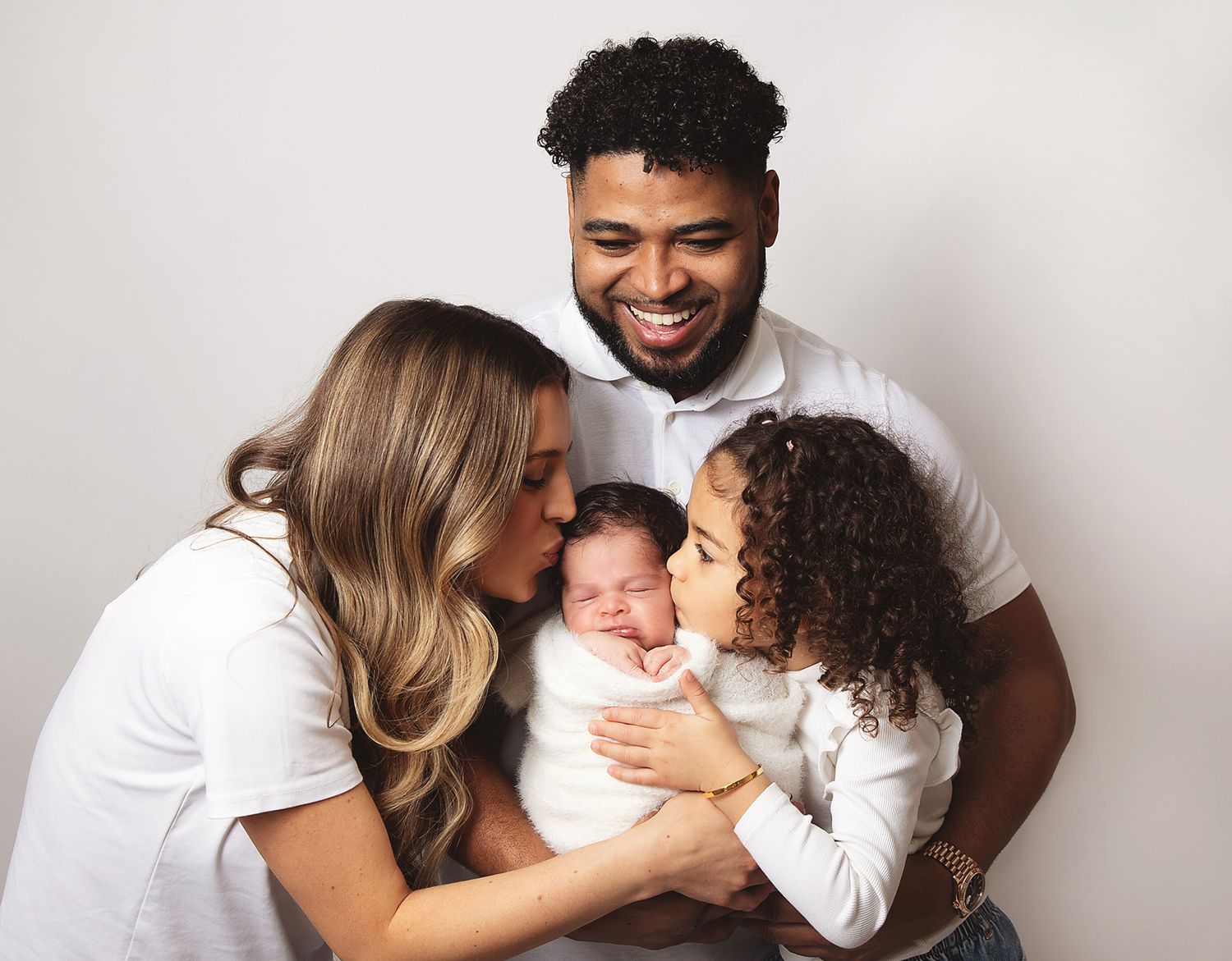 A loving family in white clothing cuddles together with their newborn baby against a light grey backdrop. During a newborn family session.