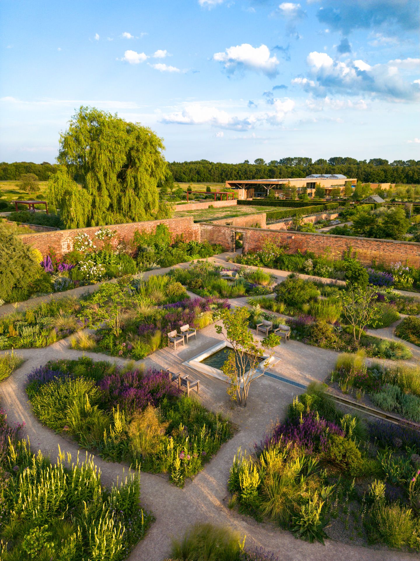An aerial view shows a beautifully landscaped garden with winding paths, colorful flower beds, and willow trees in summer.