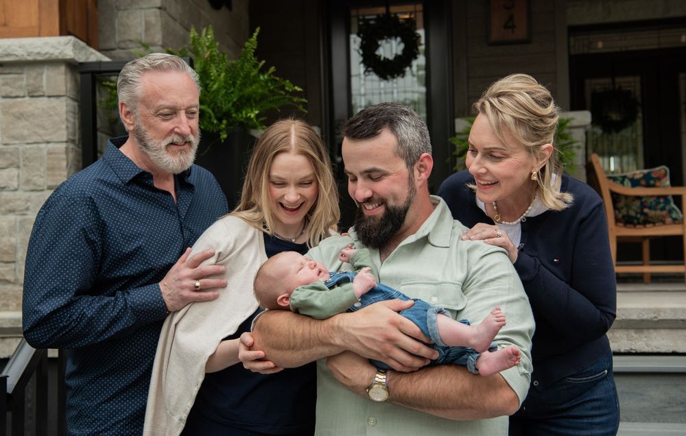 Group of adults smiling and looking down at a newborn baby wrapped in a blanket on a front porch.