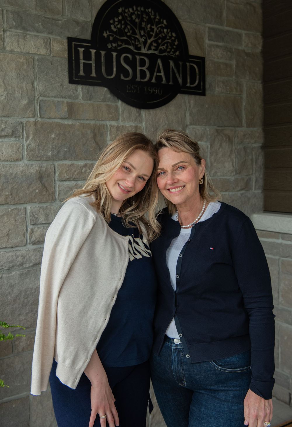 Two people stand smiling together under a decorative wall sign that reads 'HUSBAND' on a stone brick wall.