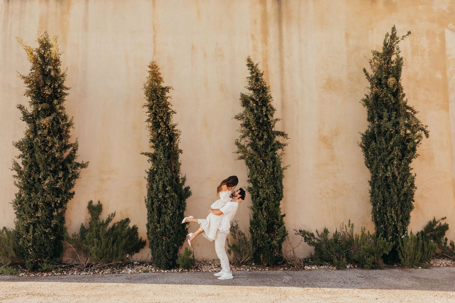 A couple embraces against a beige wall with four tall cypress trees creating an elegant outdoor backdrop.