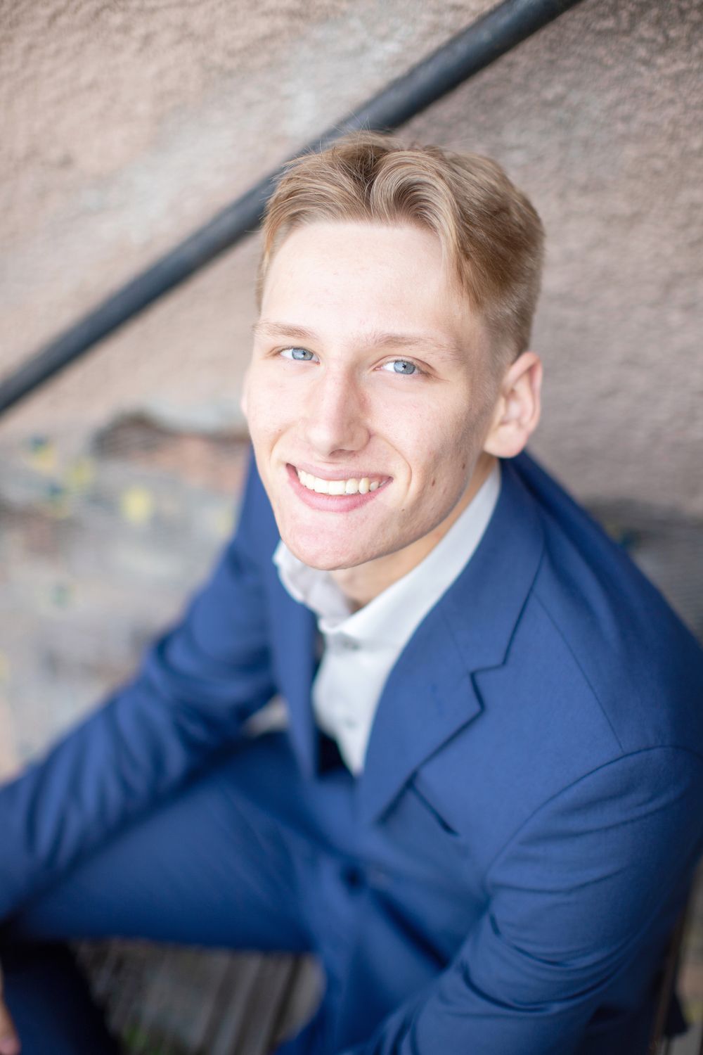 Professional headshot of someone wearing a navy blue suit jacket and light colored shirt sitting against a textured wall.