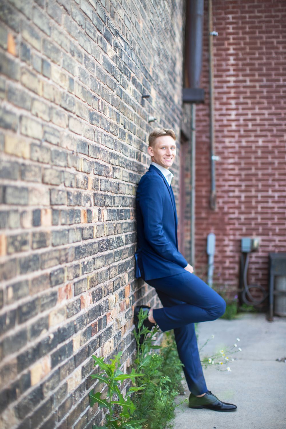 Someone in a navy blue suit poses casually against a brick wall in an urban setting.