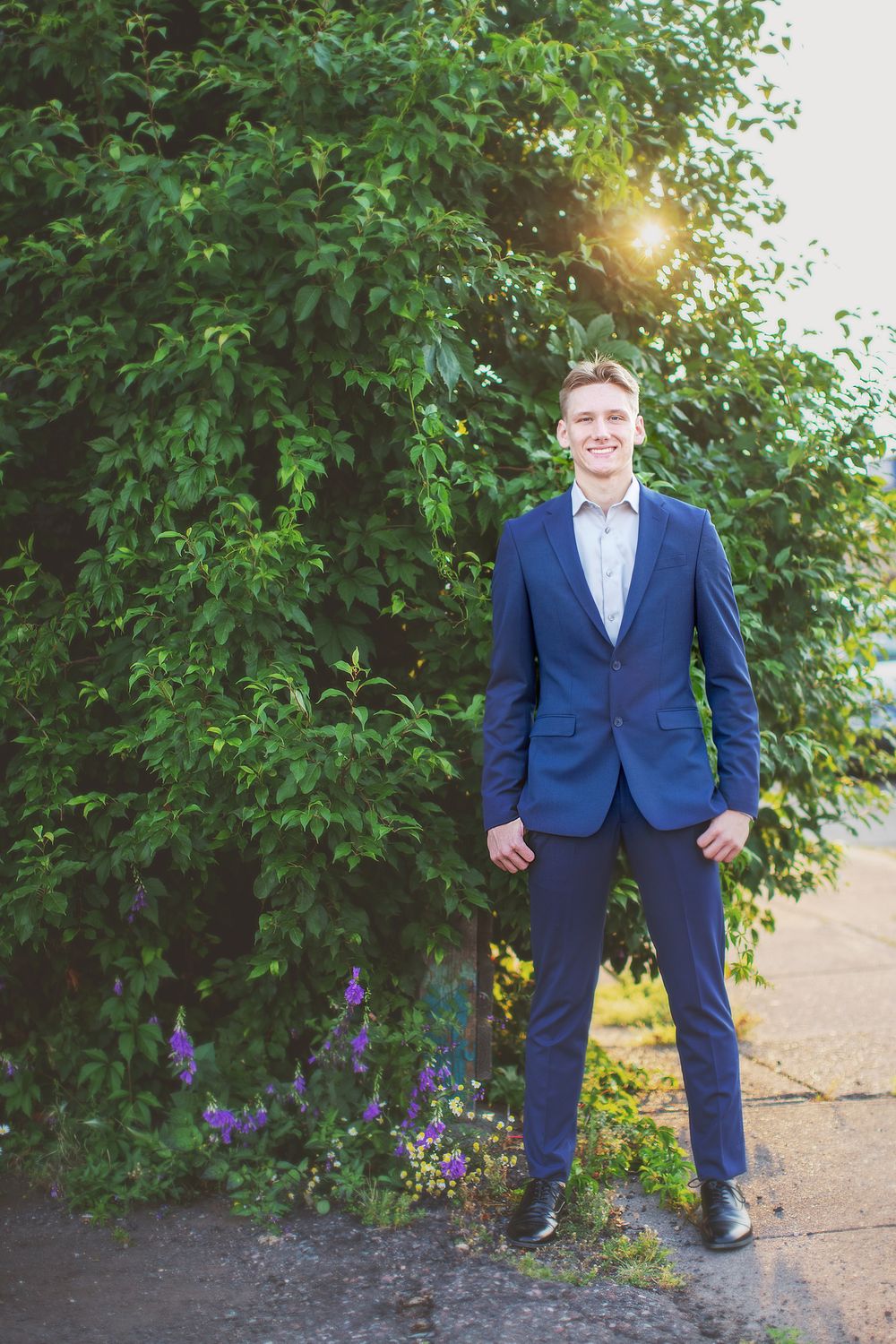 Person in a navy blue suit stands confidently by green foliage and purple flowers with sunlight peeking through.