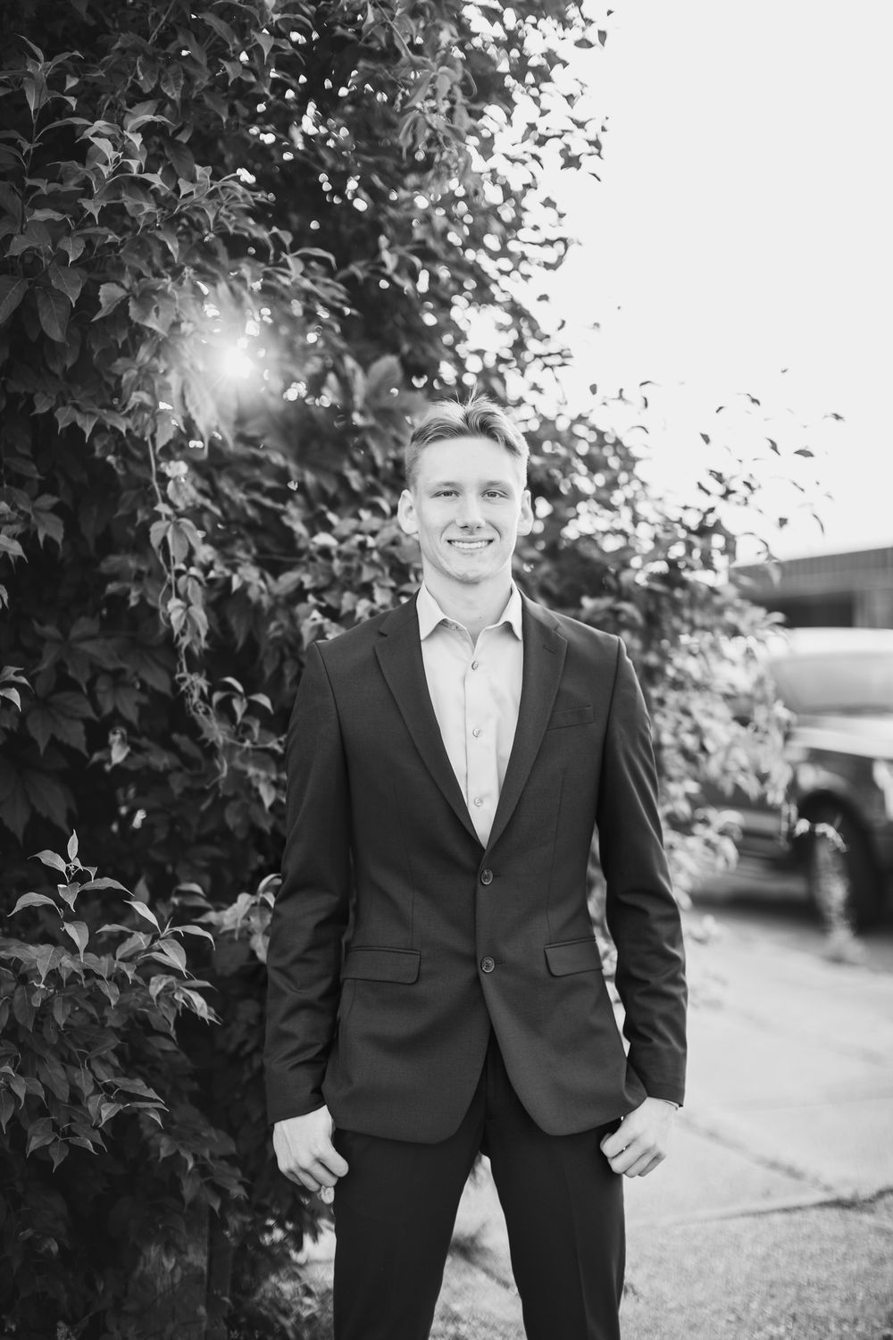 Black and white portrait of a person in a dark suit and dress shirt standing outdoors next to foliage on a sunny day.