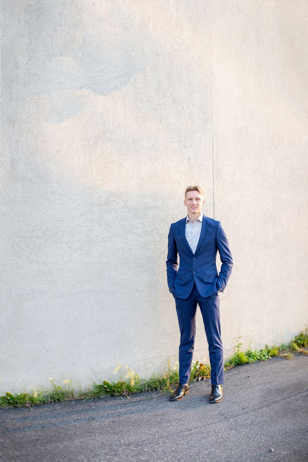 A person in a tailored navy blue suit poses against a light gray wall on a sloping path with grass peeking through.