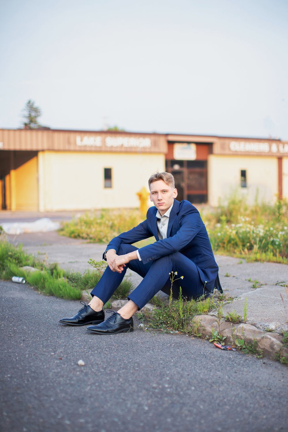 Person in navy suit sitting casually on asphalt outside a commercial building on a sunny day.
