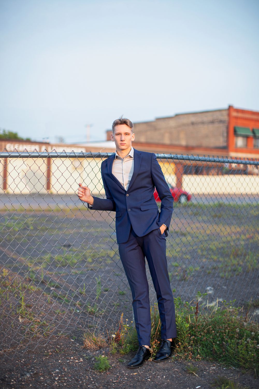 Professional wearing a navy suit poses casually near a chain link fence in an urban outdoor setting at sunset.
