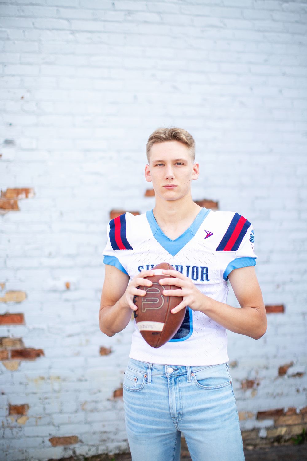 High school athlete in white jersey and jeans poses against brick wall holding football for senior portraits in triptych.