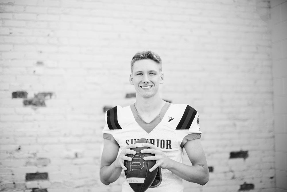 Black and white portrait of high school football player wearing jersey and holding football with white brick wall background.
