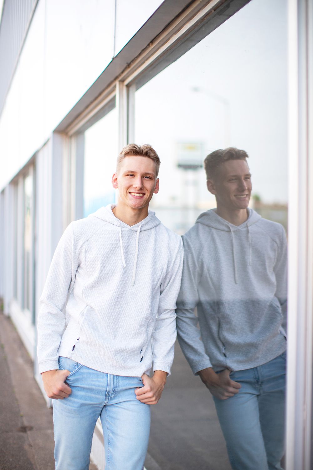 Person wearing a white hoodie and light blue jeans leans against reflective window in urban setting.