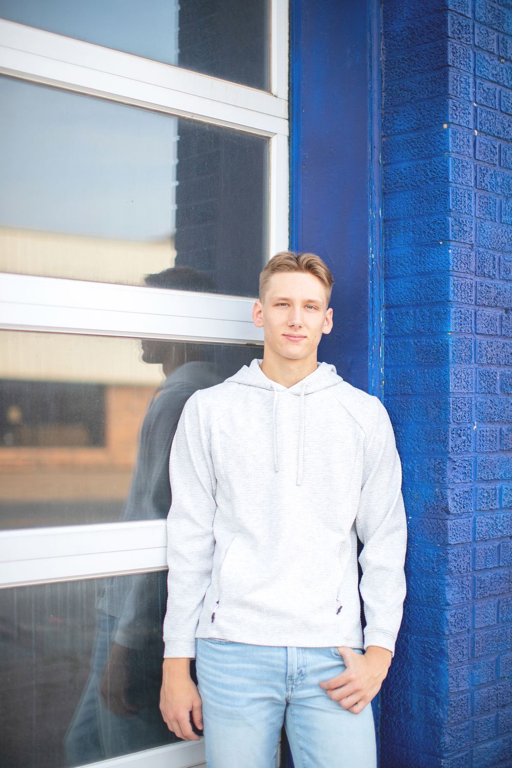 Person in light grey hoodie and jeans standing against bright blue wall and large window.