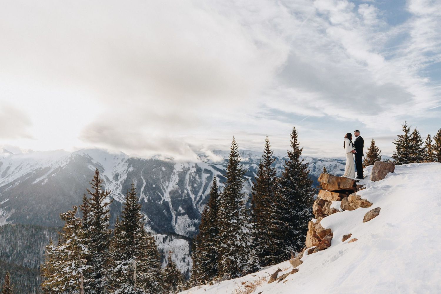 Two people stand on a snowy mountain overlook with evergreen trees and dramatic mountain peaks in the background.