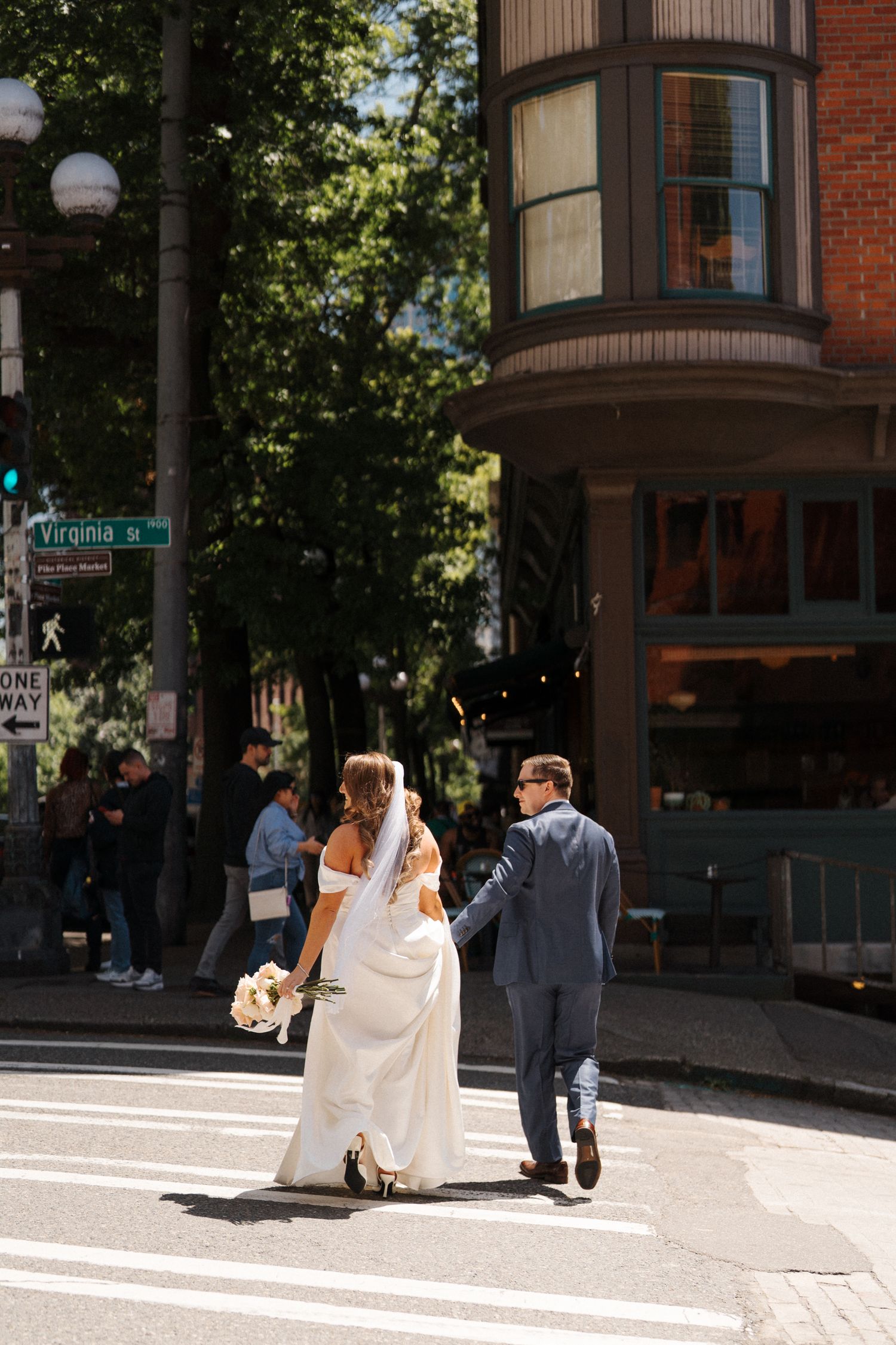 A couple in wedding attire walking across a city street on a sunny day near a brick building with a bay window.