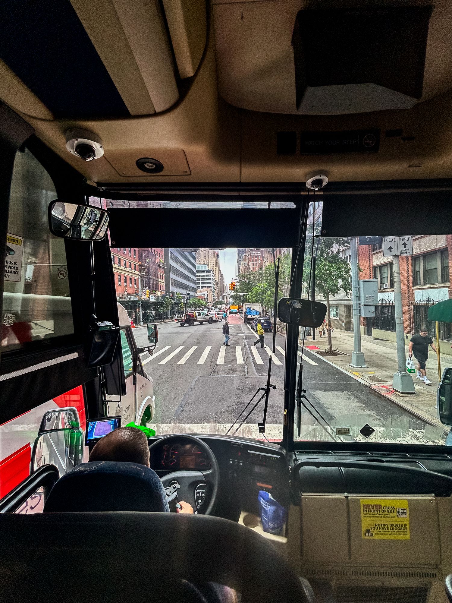 View from bus driver's seat looking out at busy city street intersection with crosswalk and buildings in New York City.