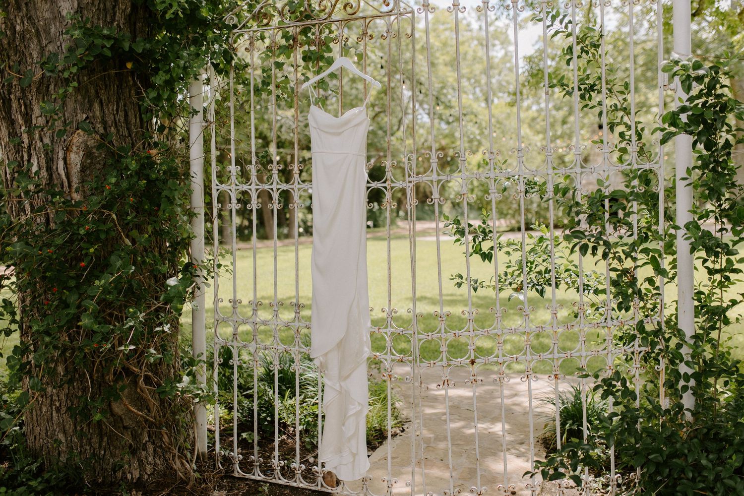 A white garden gate covered in climbing vines opens to a sunlit garden path.