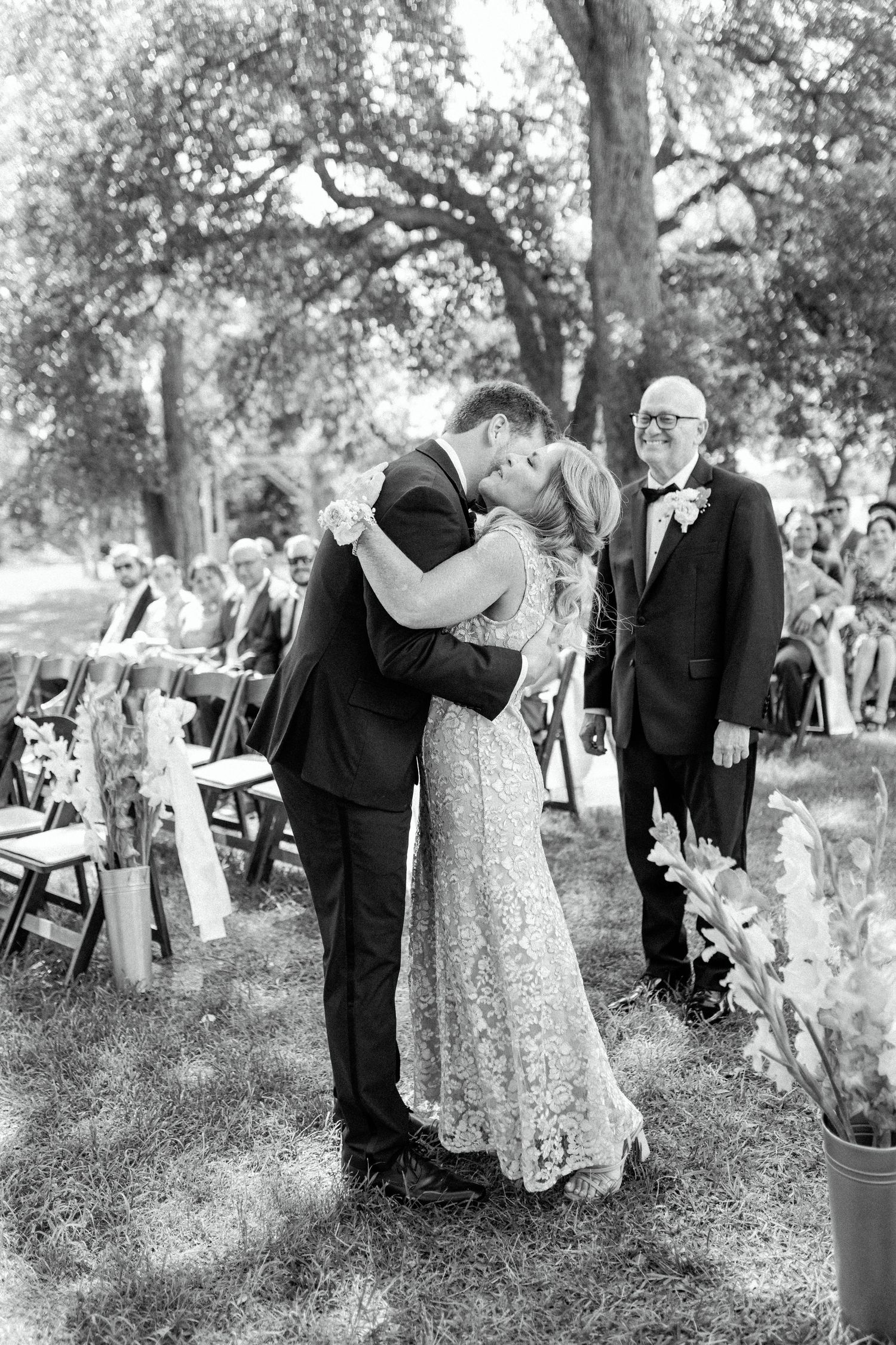 Black and white outdoor wedding ceremony photo with couple sharing first kiss under trees.
