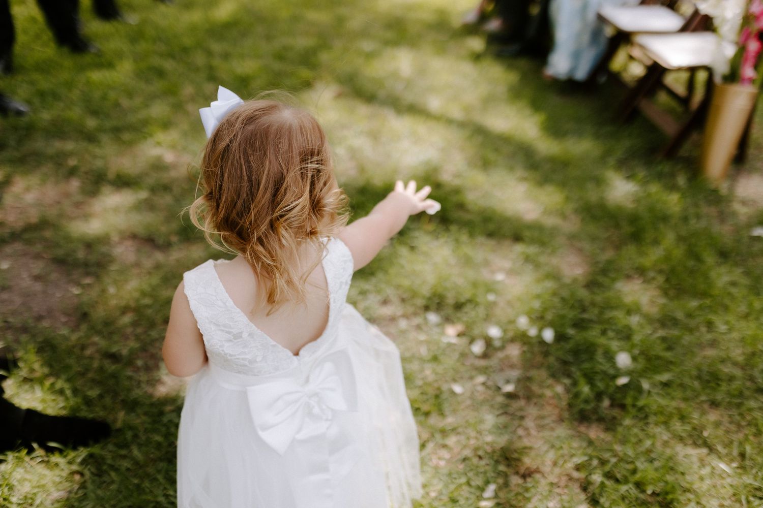 A young flower girl in white dress gestures while standing on a grassy outdoor wedding venue path.