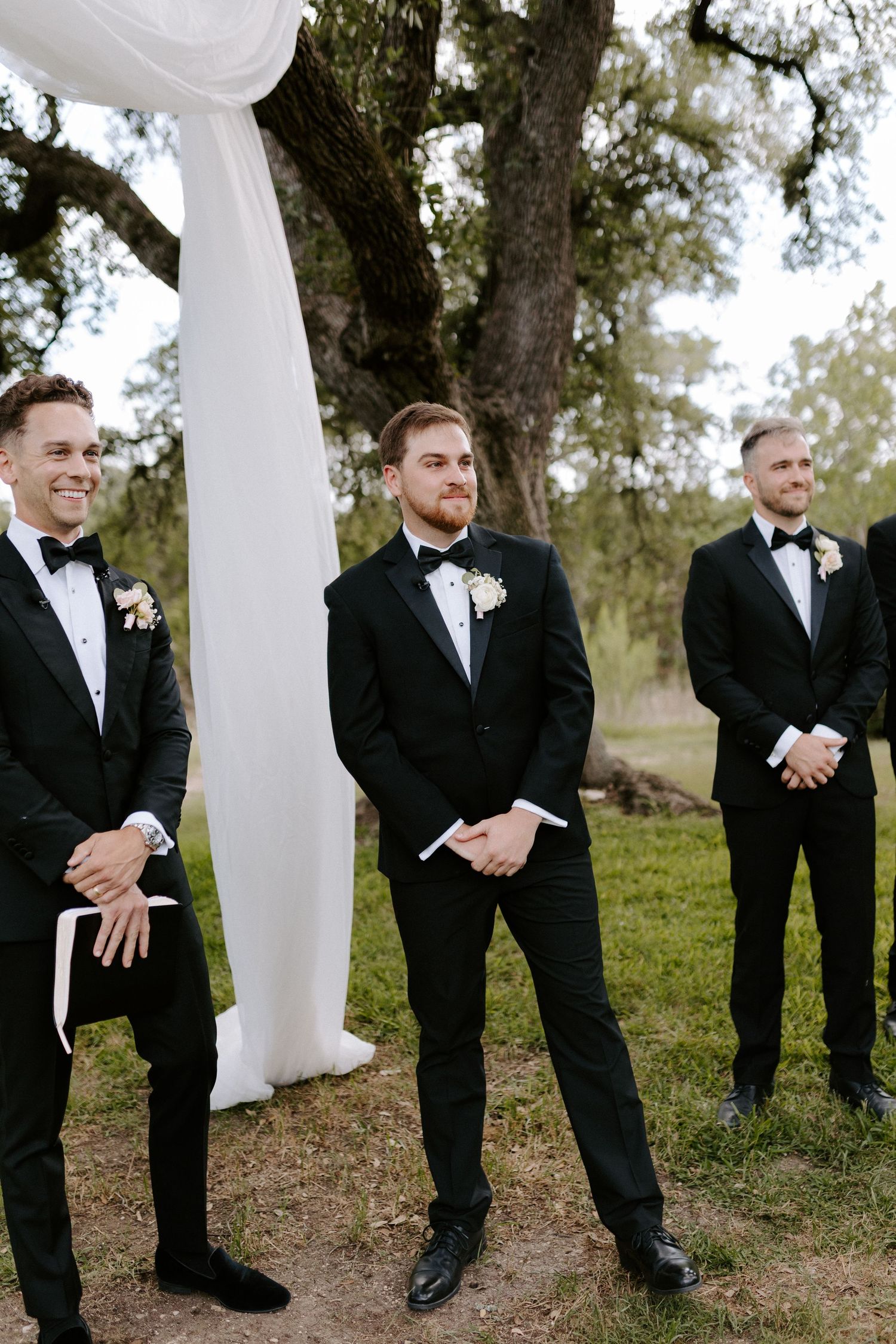 Three groomsmen in black tuxedos stand against a white draped backdrop outdoors.