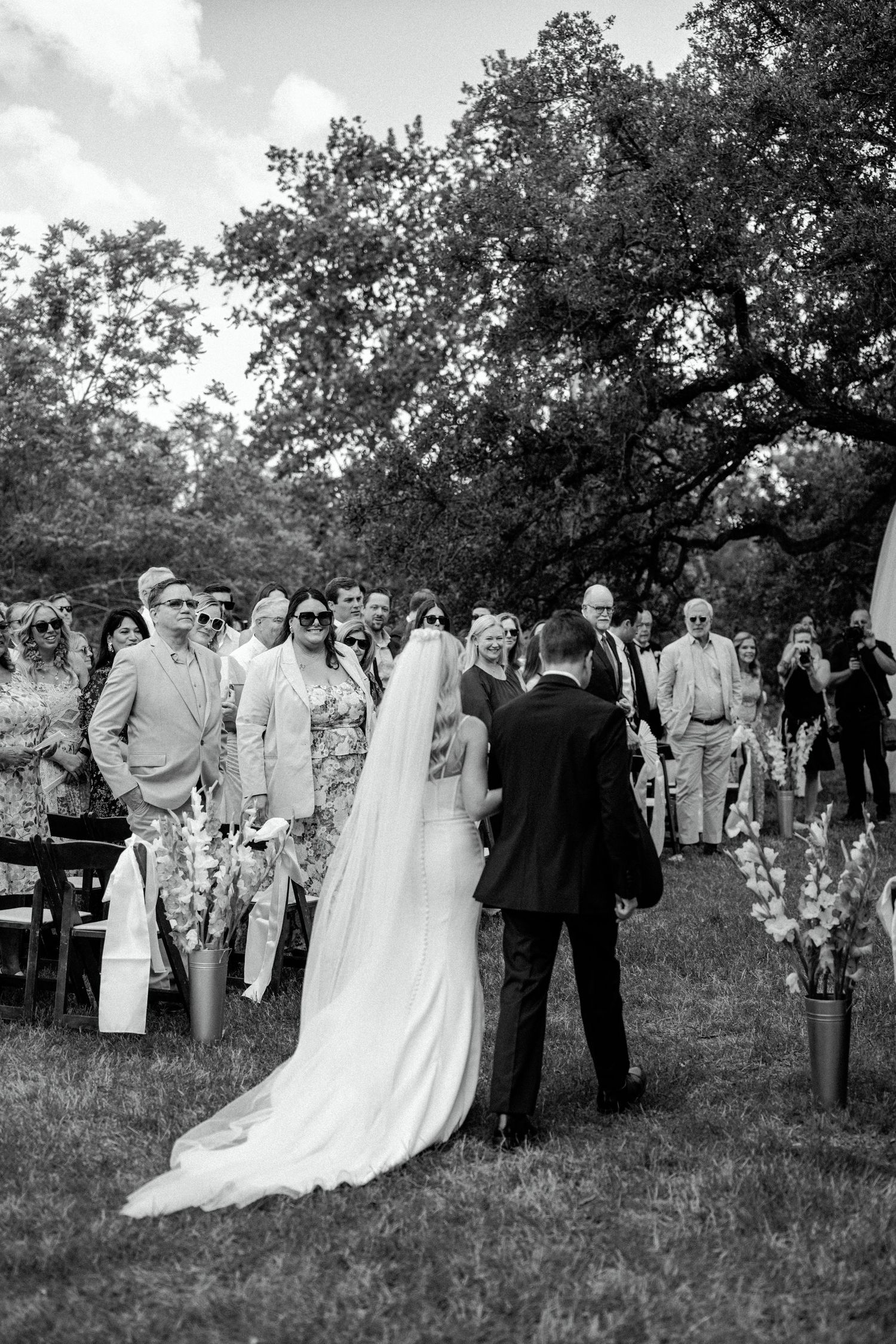 Black and white photo of an outdoor wedding ceremony under large trees.