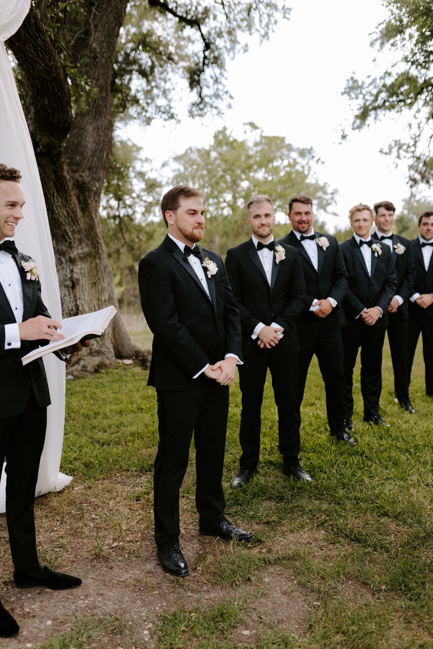 A group of groomsmen in black tuxedos line up outdoors under trees at a wedding celebration.