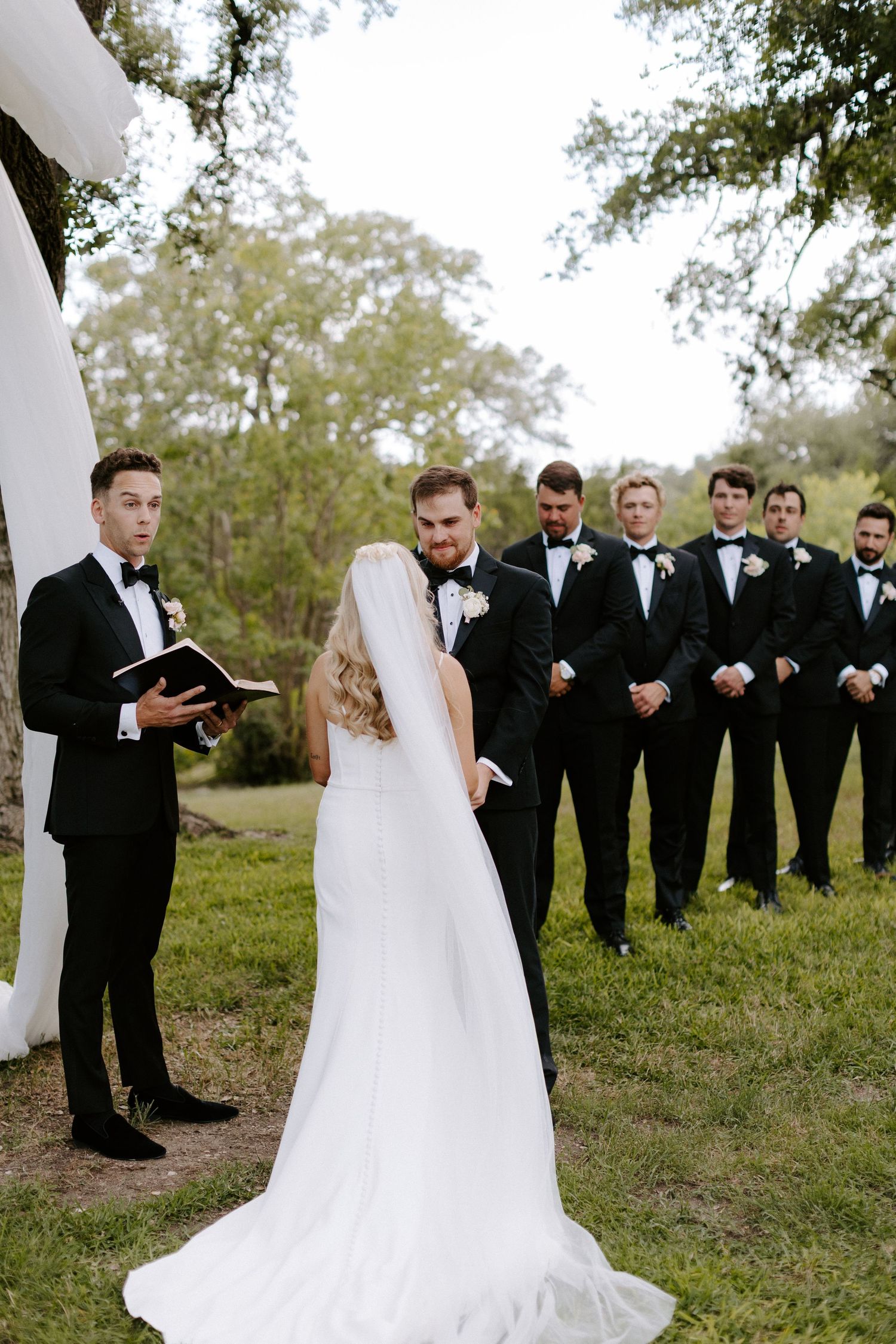 Wedding party gathered on grassy hillside with bridesmaids in black suits and white gown.
