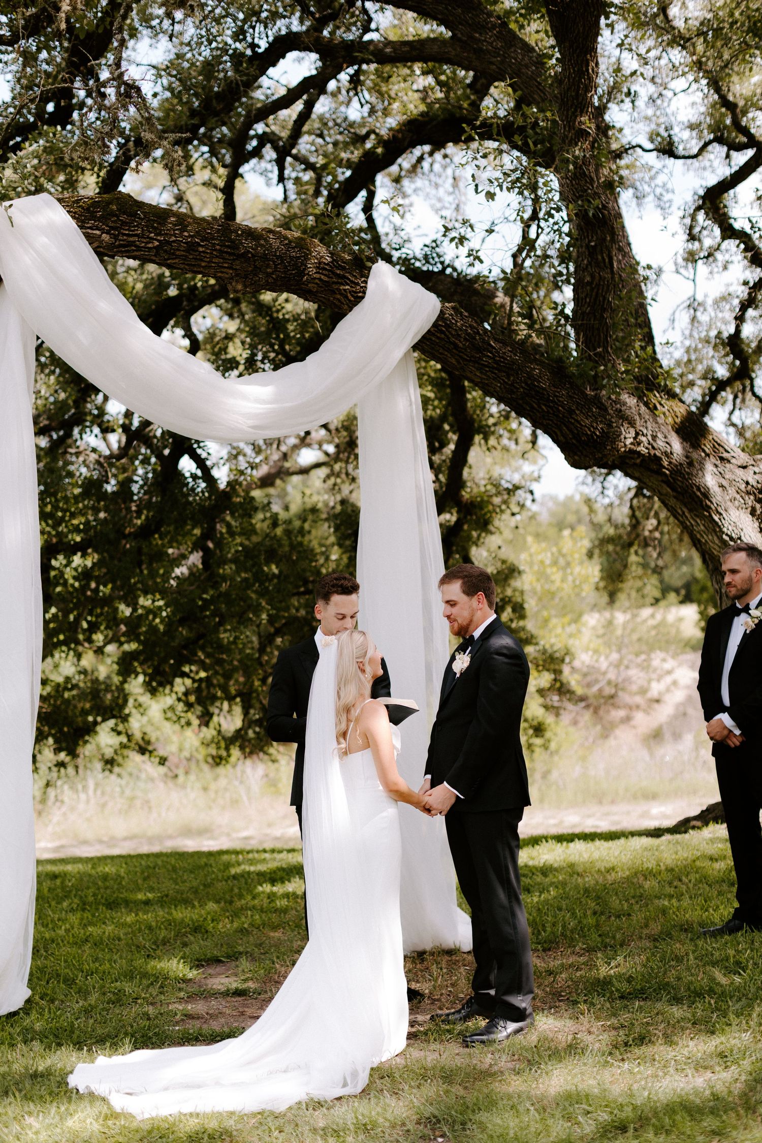 Romantic outdoor wedding ceremony with white fabric draped between trees creating an elegant natural backdrop in sunlight.