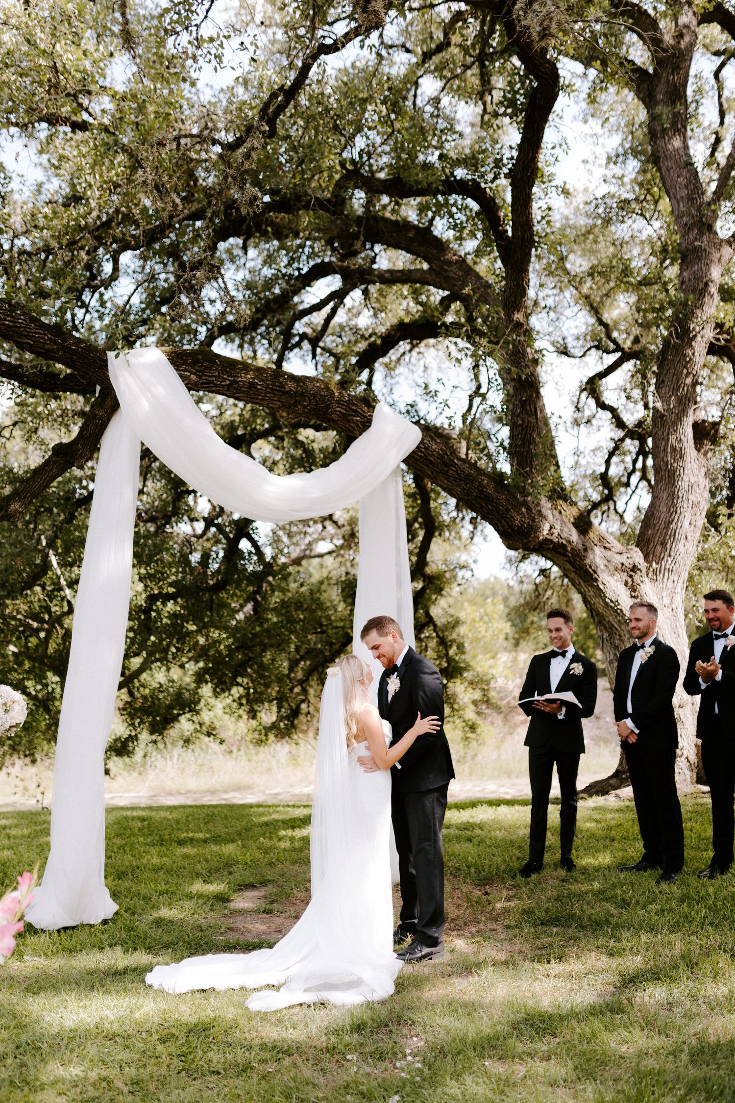 Outdoor wedding ceremony under draped white arch with oak trees in background.