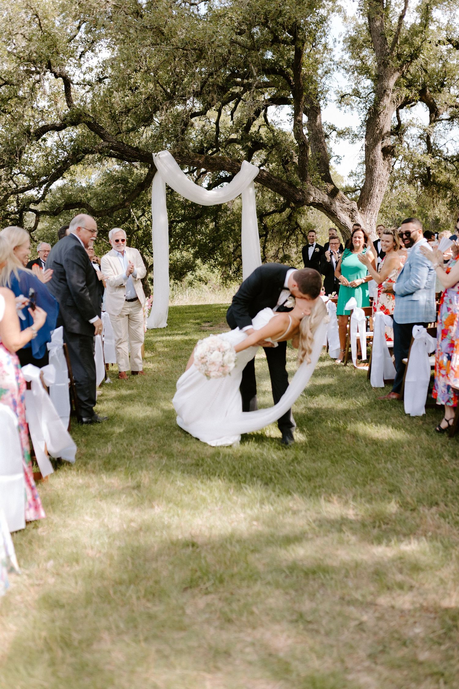 Wedding guests celebrating outdoors under oak trees during daytime ceremony.
