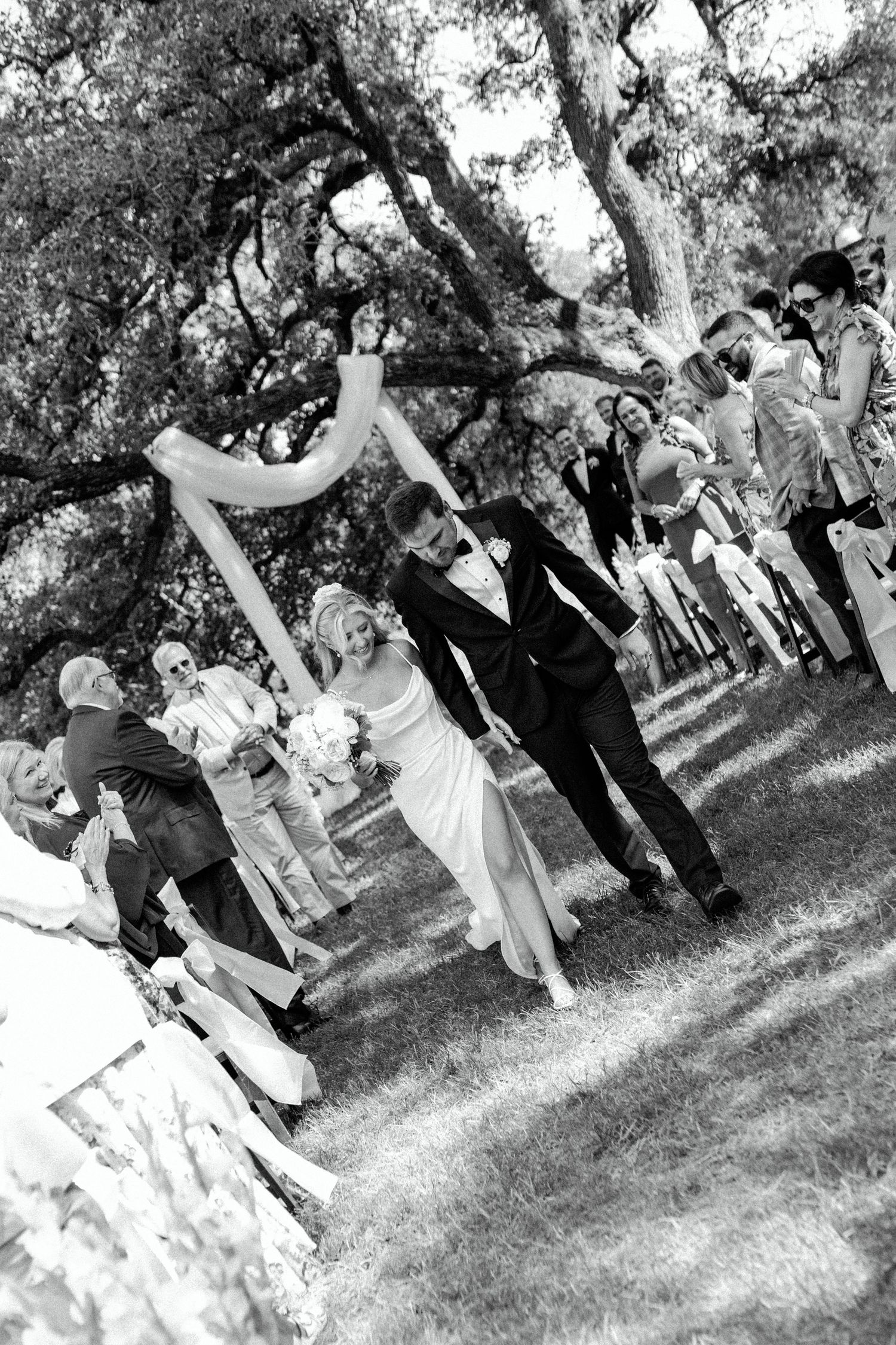 Black and white photo capturing joyful celebration at outdoor wedding ceremony under shady trees.