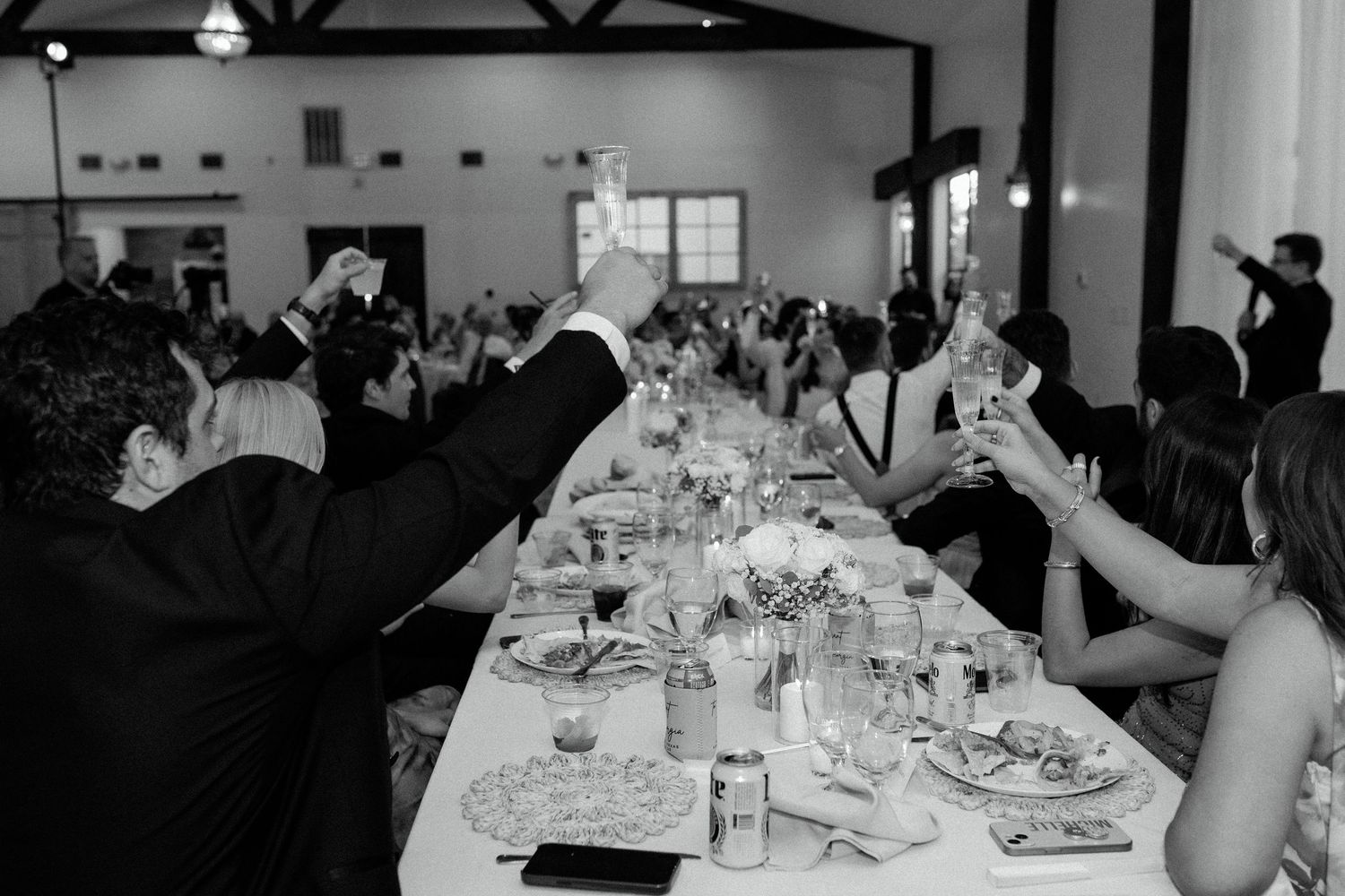 Black and white photo of wedding guests raising glasses in a toast at long reception table.