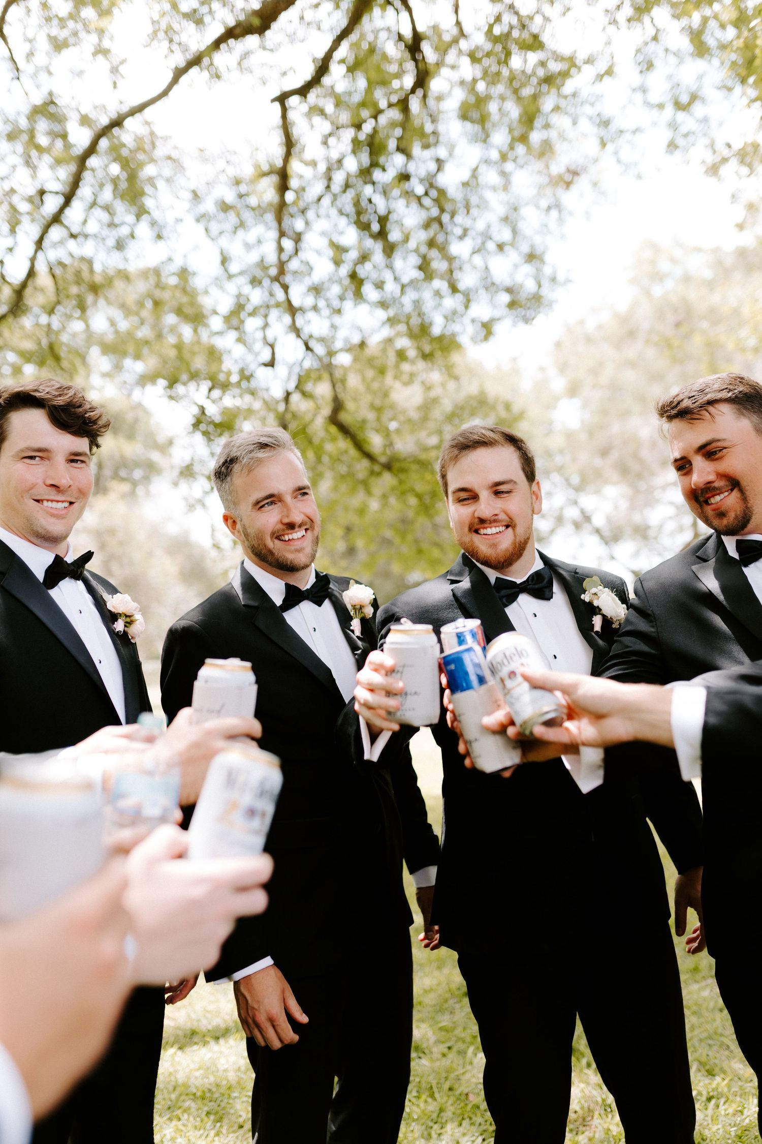Groomsmen in black tuxedos toasting with drinks outdoors under tree branches.
