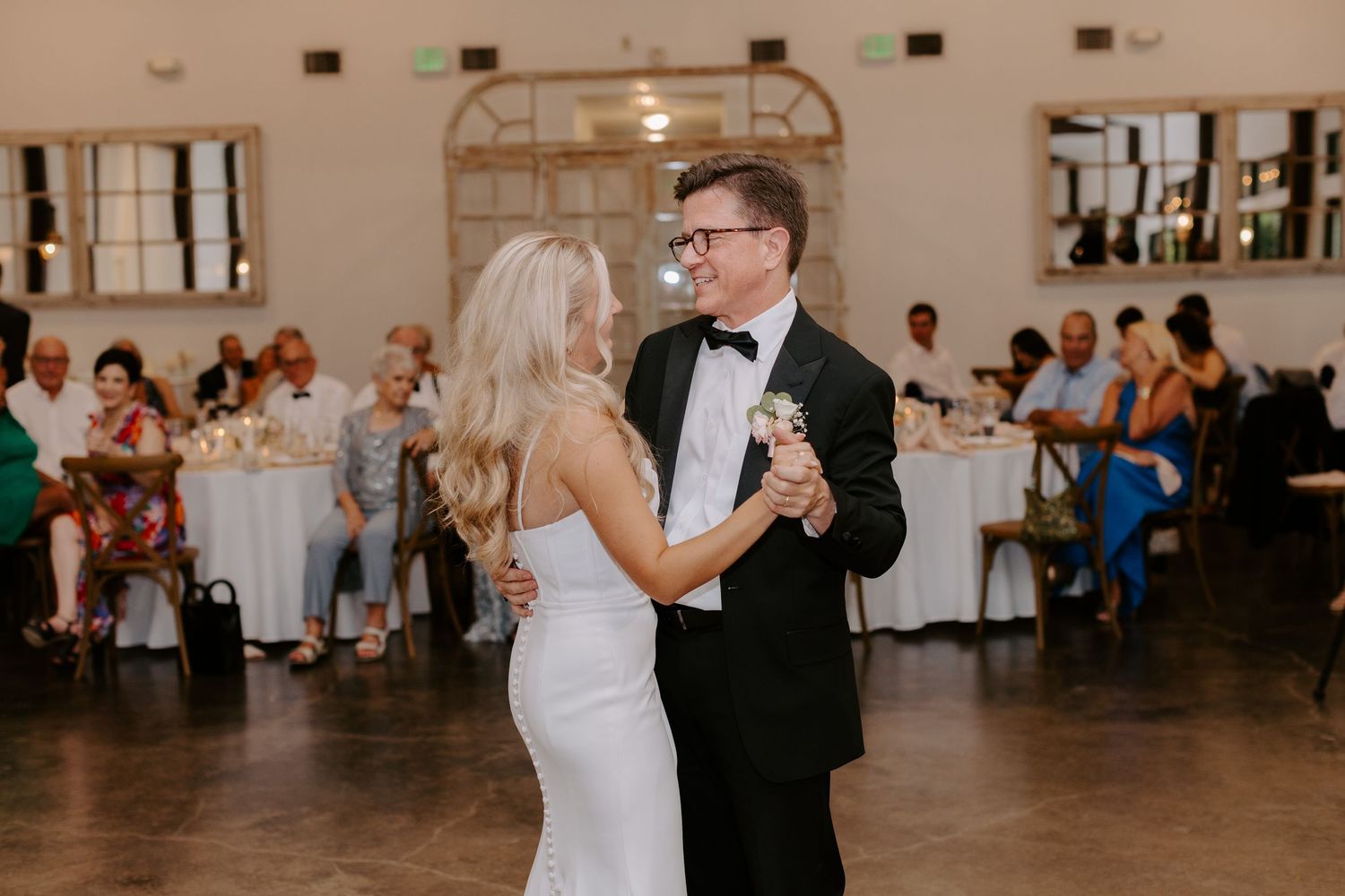 Elegant couple sharing first dance in reception hall with guests seated at tables in background.