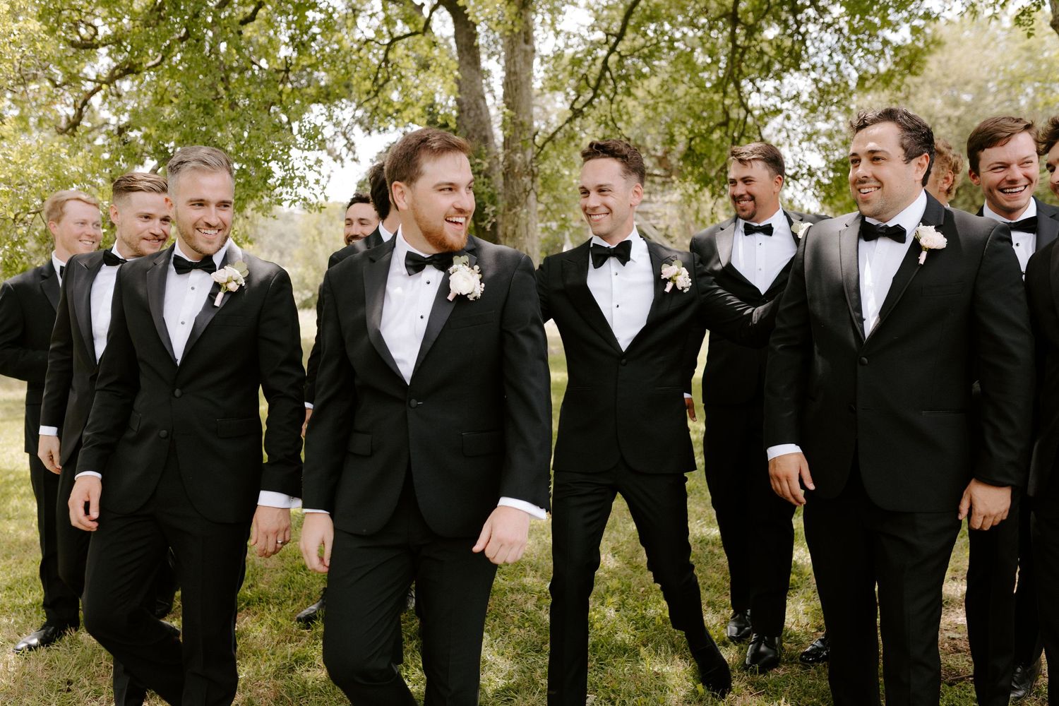 Groomsmen in black tuxedos and bow ties gathered outdoors for a wedding party photo.