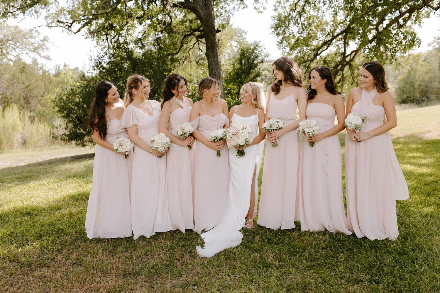 Bridesmaids in blush pink dresses pose together on sunny lawn.