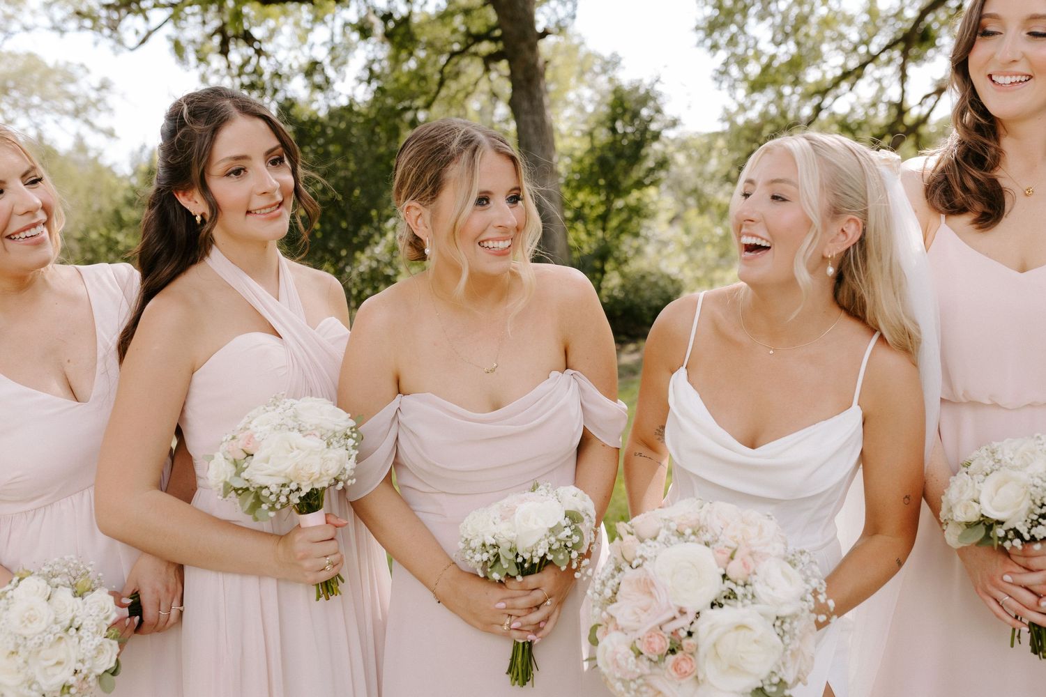Bridesmaids in soft pink dresses share joyful laughter while holding white bouquets.