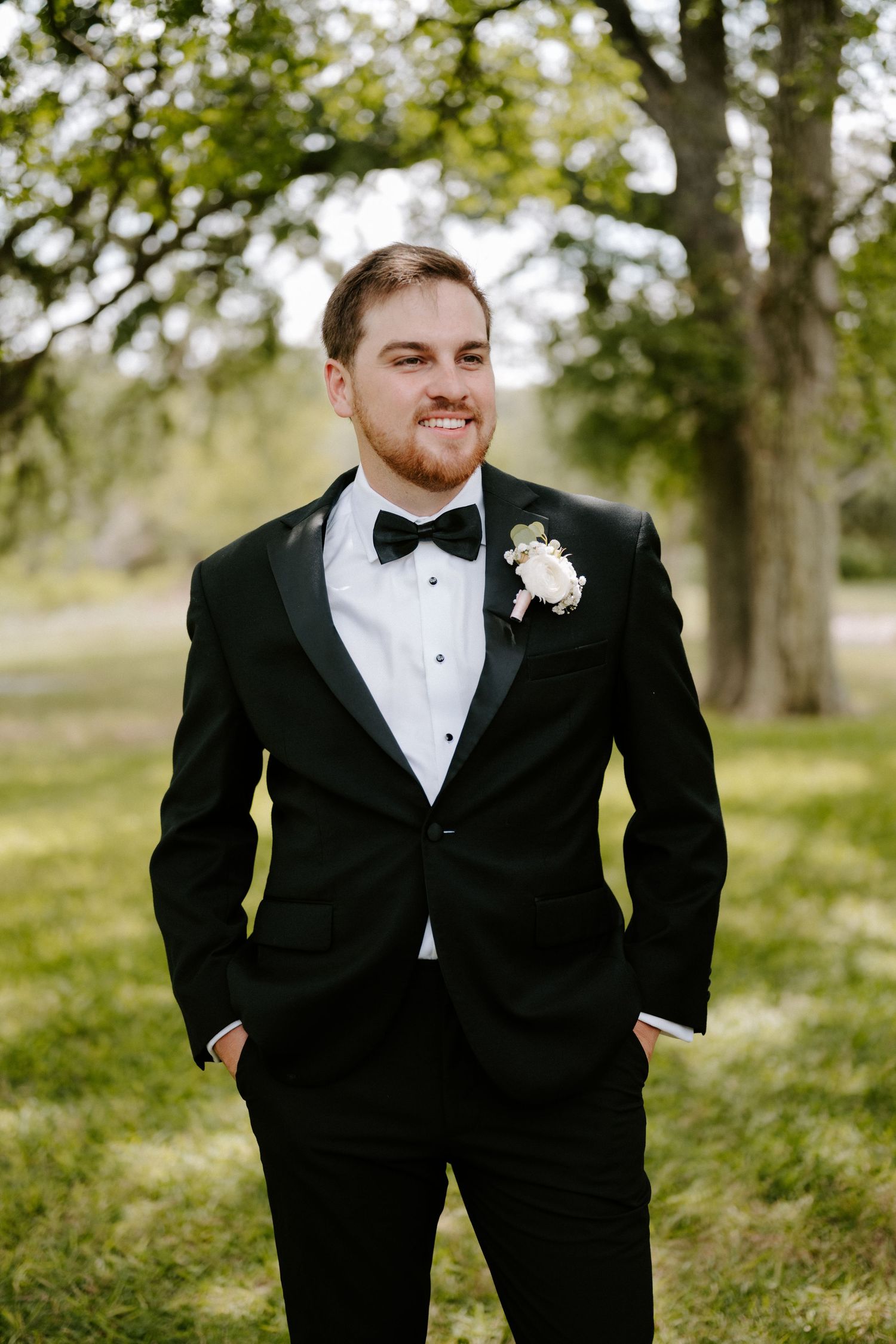 A groom in a classic black tuxedo and bow tie stands outdoors with a boutonniere against a natural backdrop.