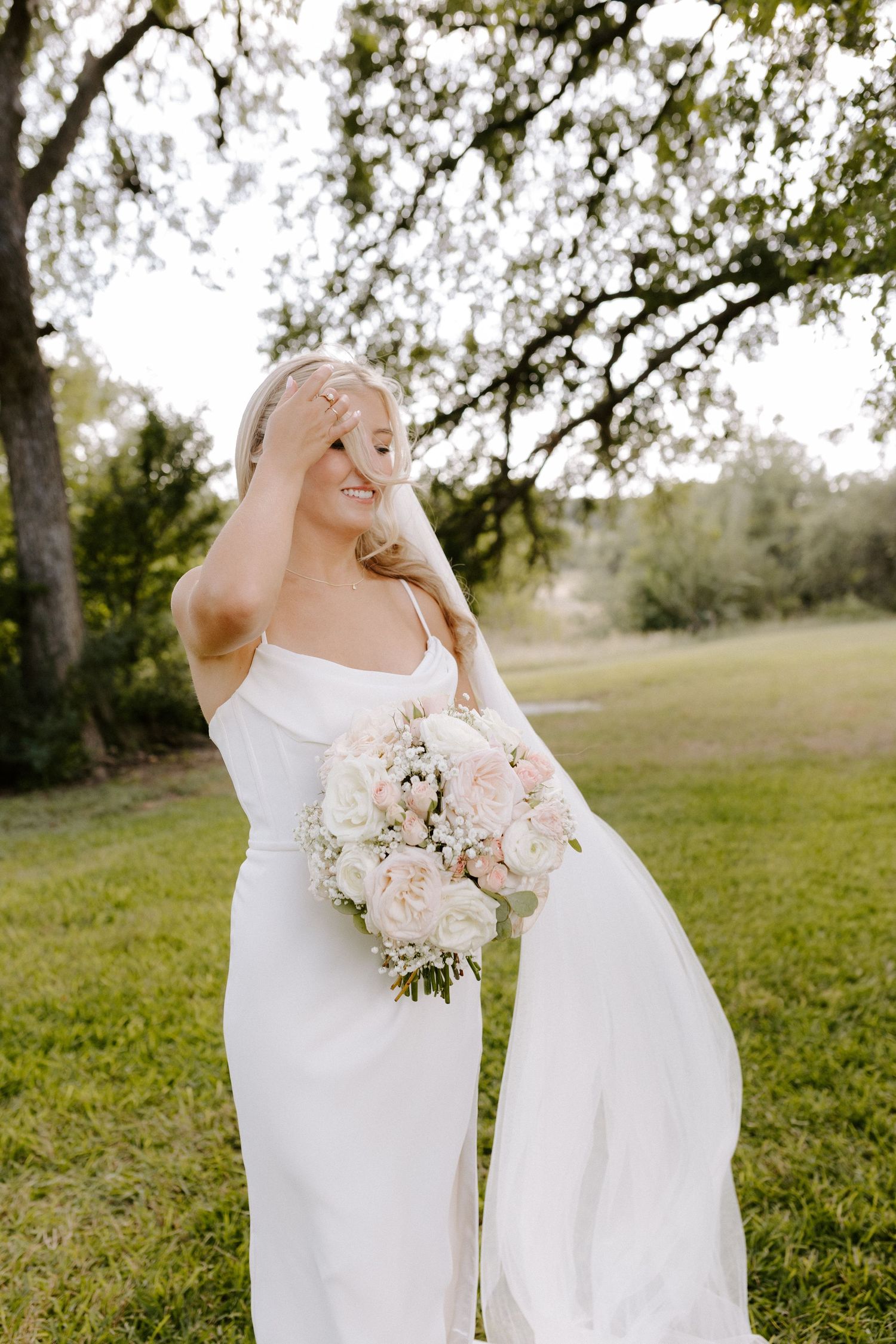 Bride in sleek white dress holds pink and white bouquet outdoors.