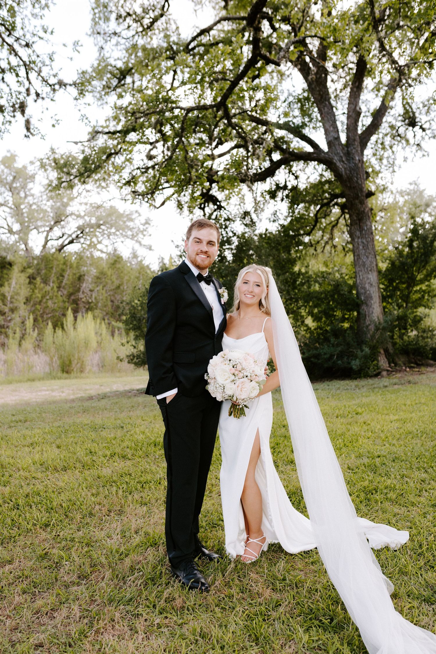 A bride in a white dress with long veil and groom in black tuxedo pose together on grassy grounds under a large oak tree.