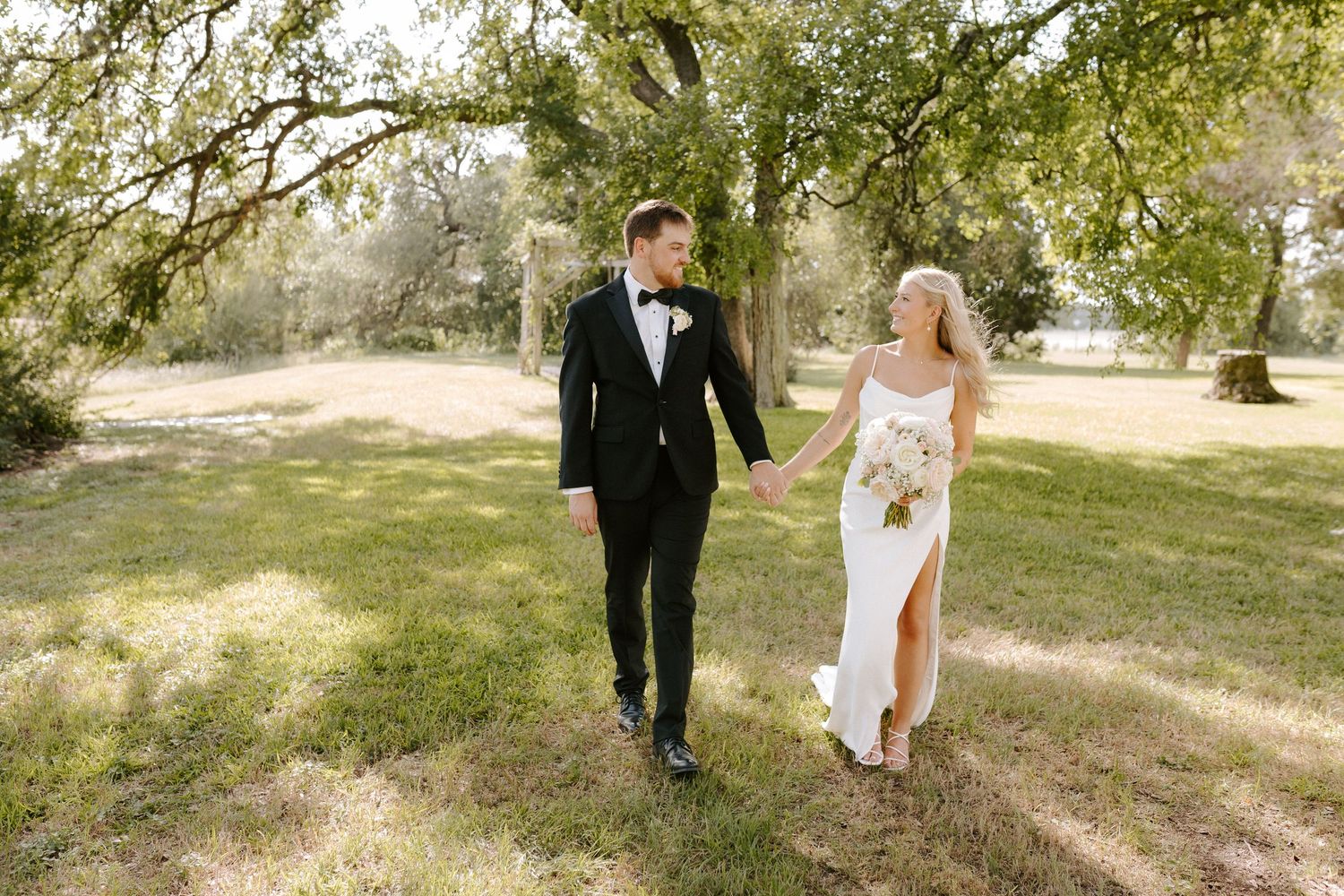 Newlyweds walking together on grass under sunlit trees.