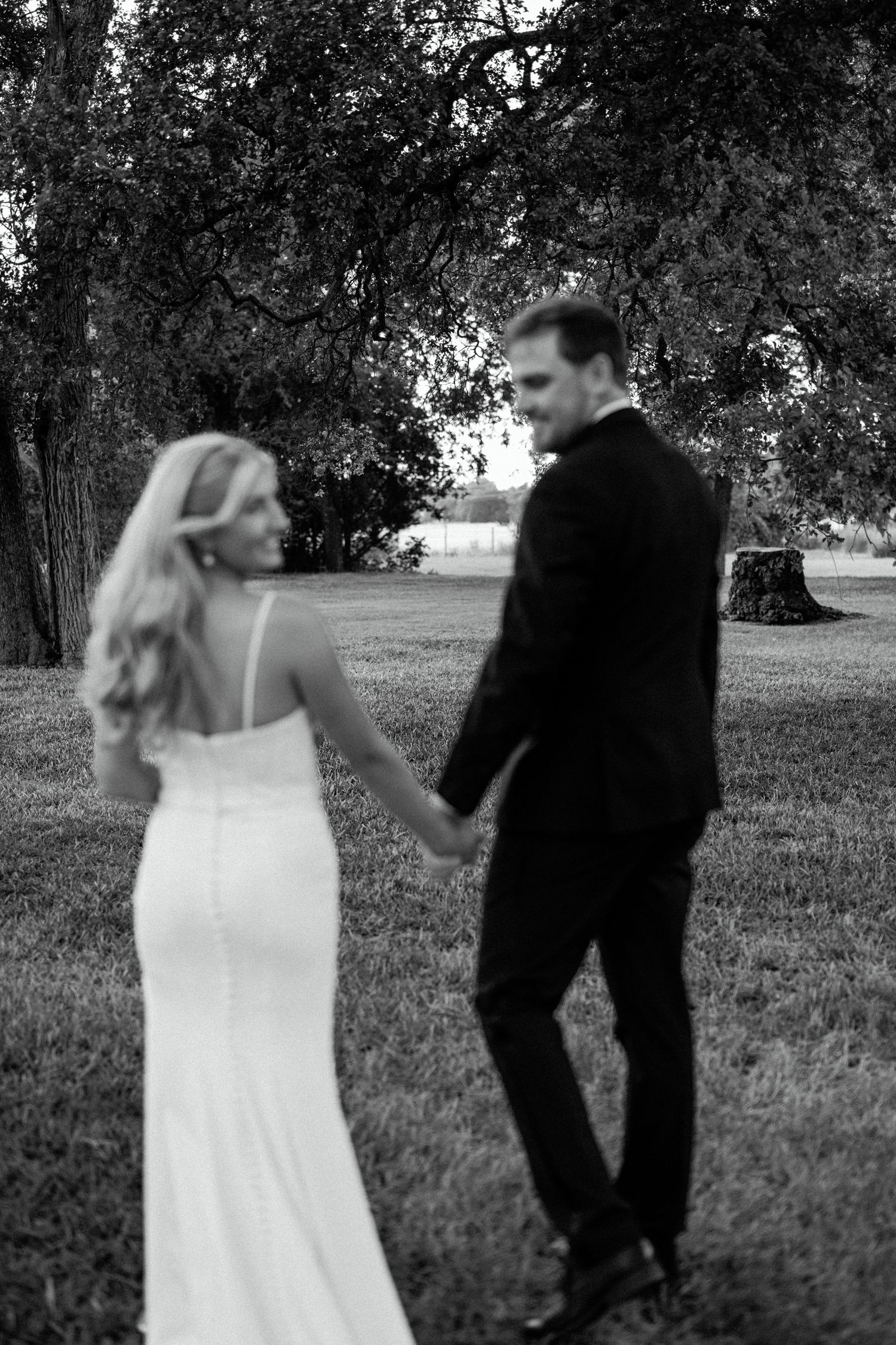 Black and white photo of a couple in formal attire holding hands outdoors on a grassy lawn.