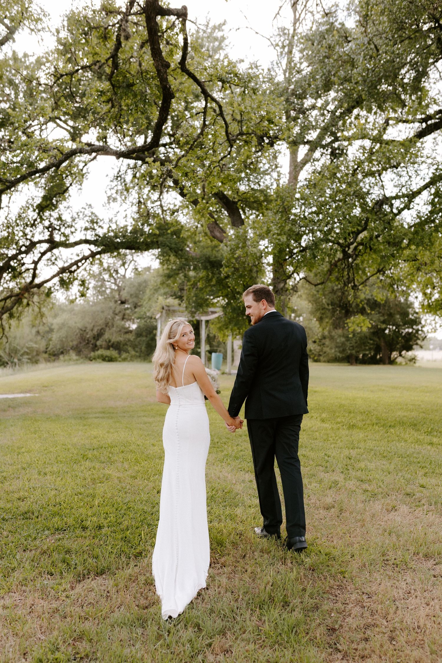 A classic wedding moment captured outdoors with a couple walking under oak trees in formal attire.