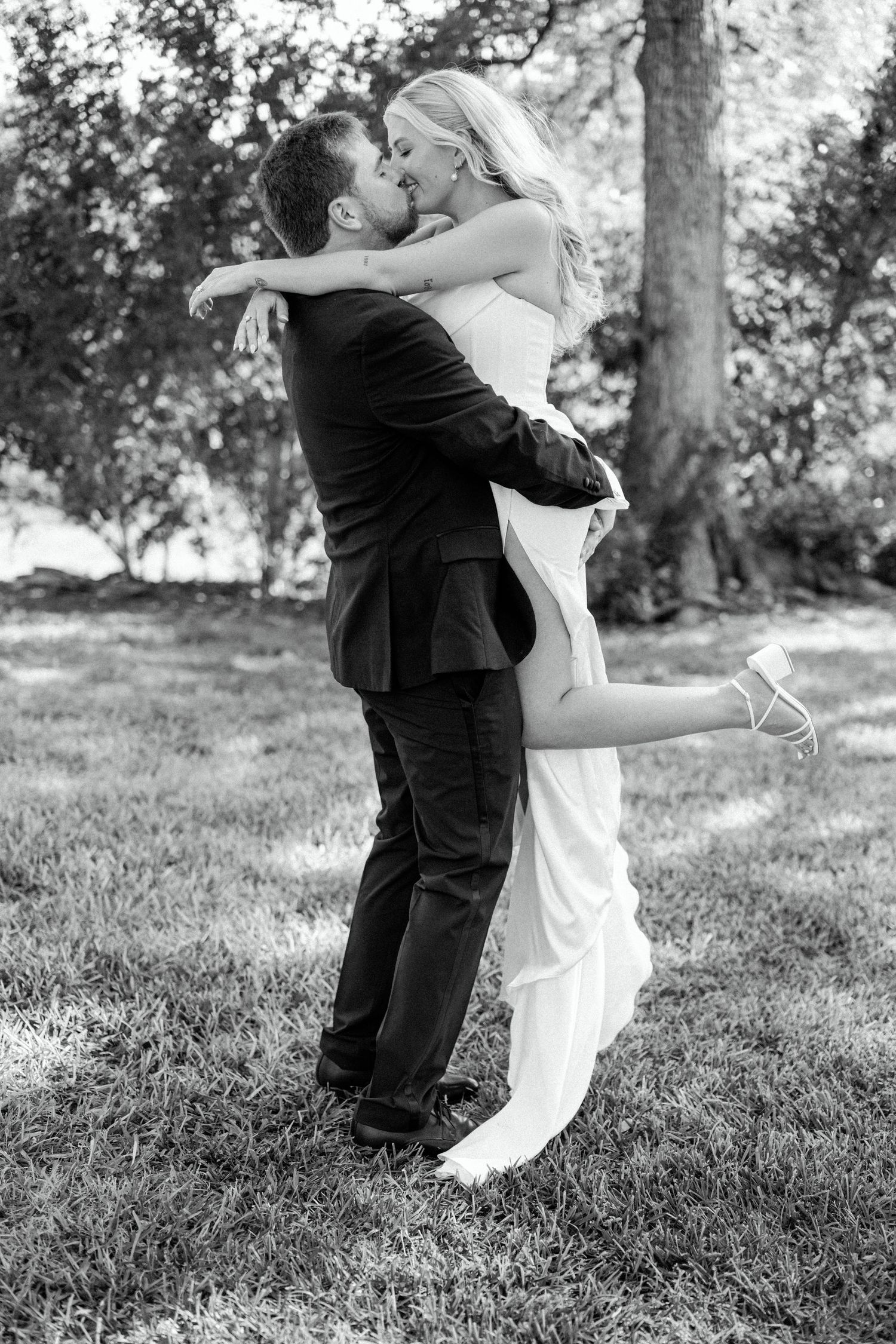 A romantic black and white wedding photo shows a couple embracing outdoors with the bride kicking up her leg.
