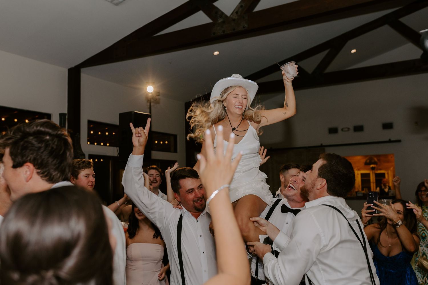 Bride in white dress being lifted up by wedding guests during lively reception celebration.