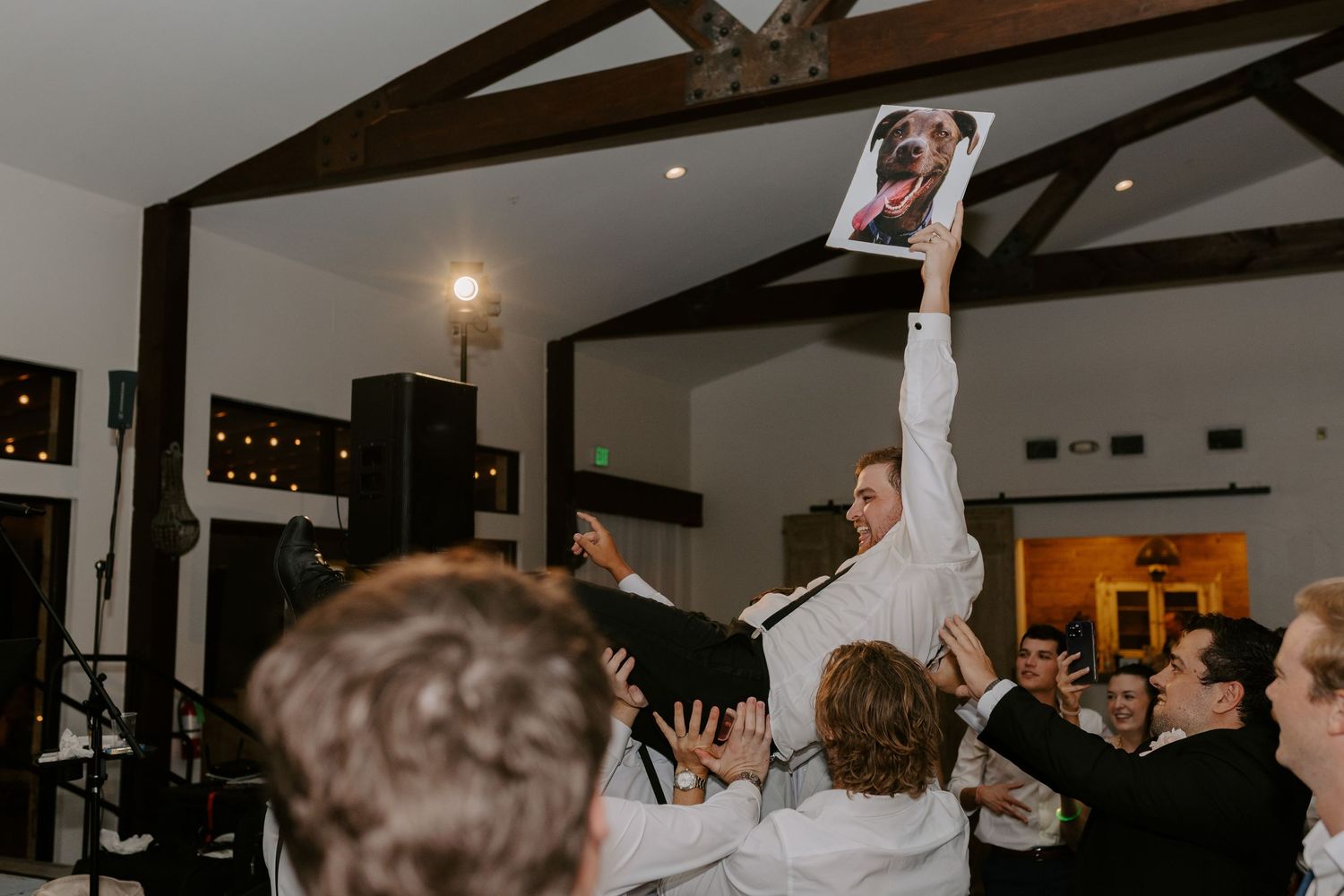 Wedding guests dancing and celebrating at a reception under wooden beam ceilings in a festive venue.