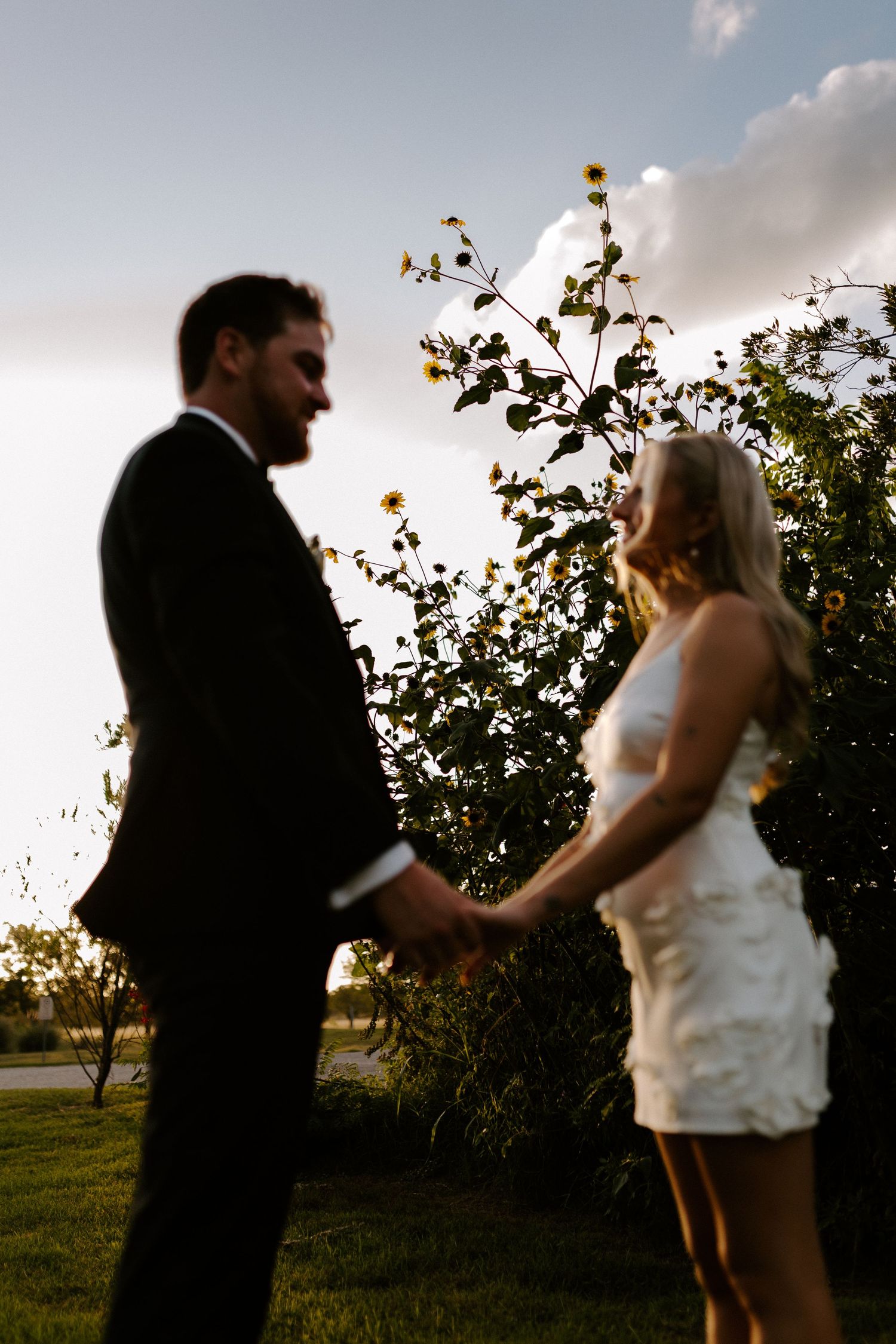 Couple in wedding attire sharing an intimate moment at sunset among flowering bushes in an outdoor garden setting.