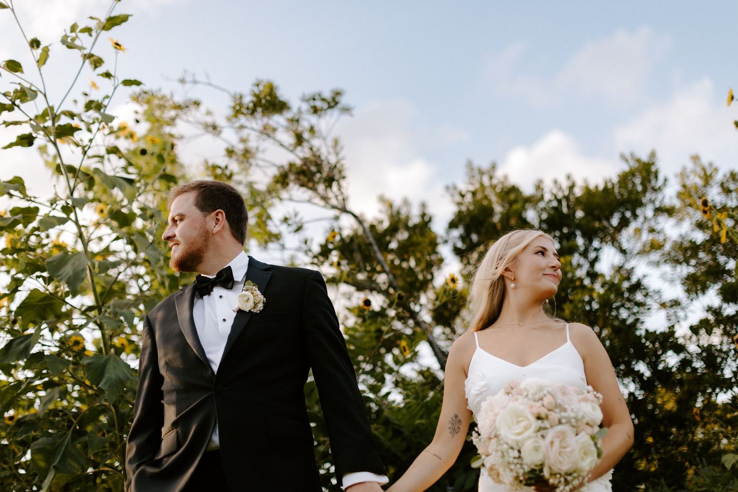 Wedding couple walking together through garden setting at sunset with white floral bouquet.