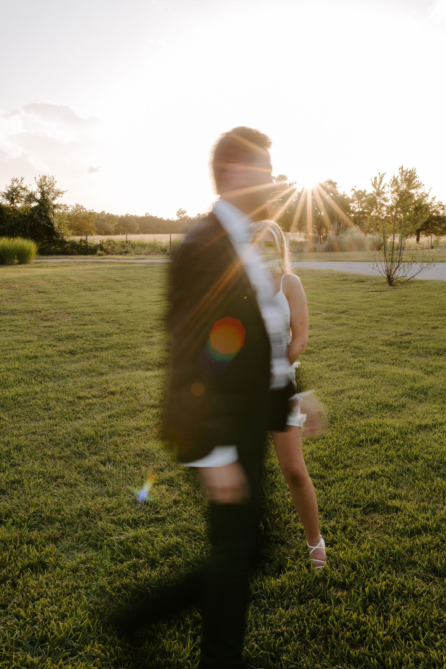 Artistic wedding photo with sun flare capturing a couple in black and white formal attire at sunset.