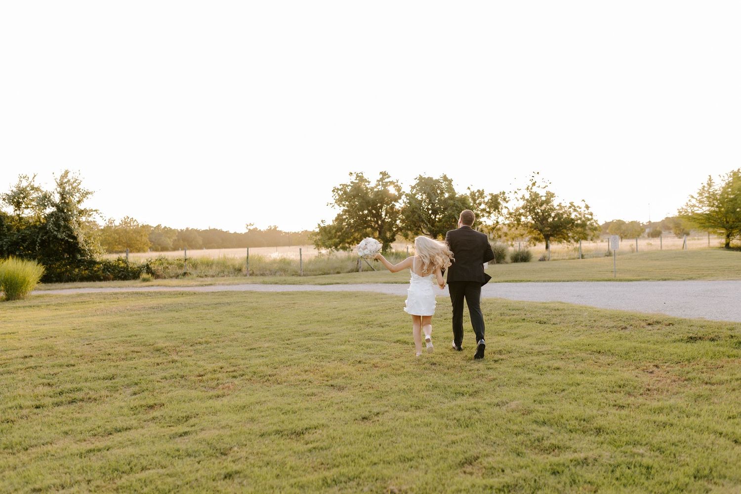 Sunset wedding portrait of couple walking together on grassy field with trees in background.