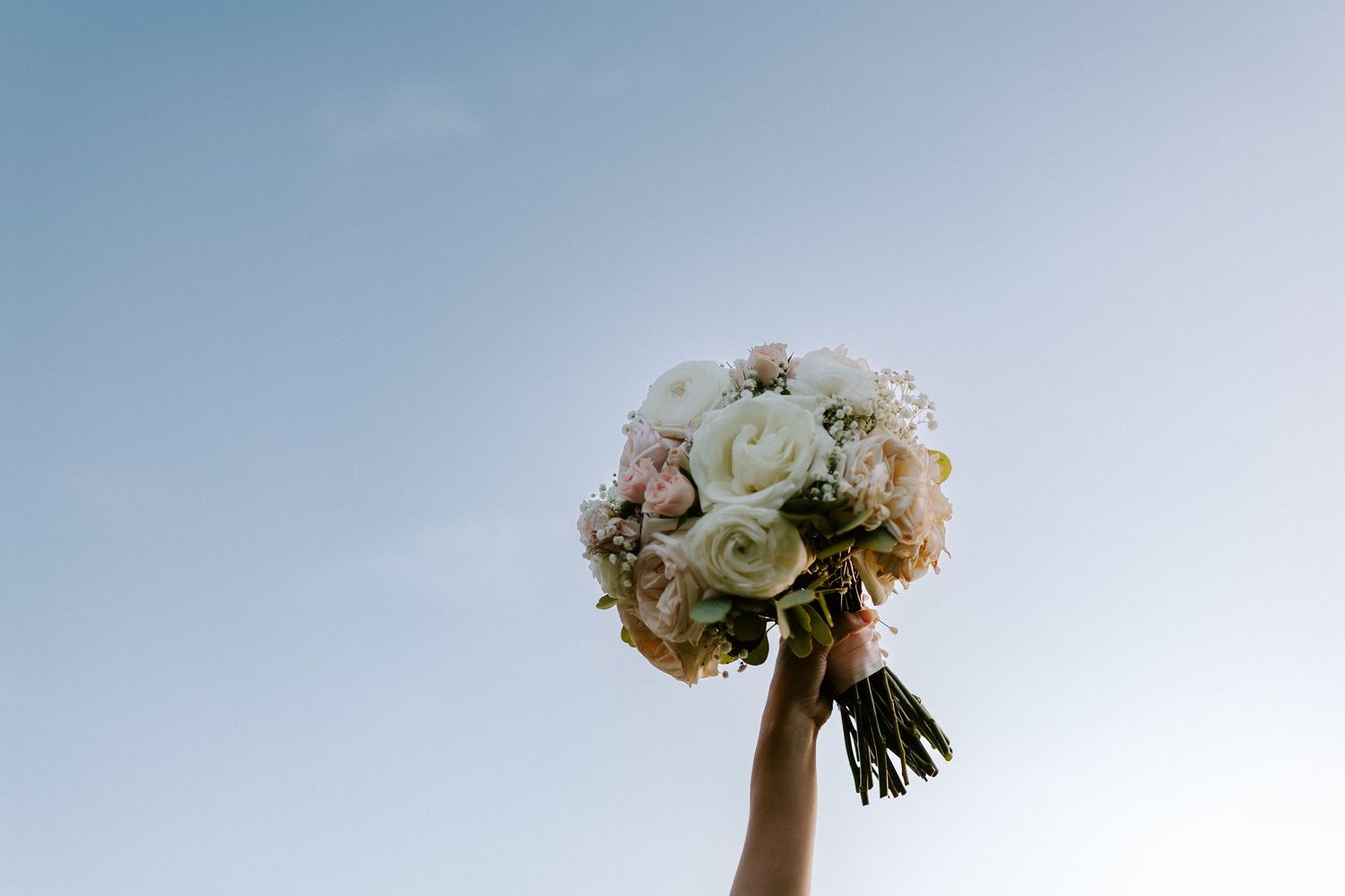White and blush wedding bouquet held up against a bright blue sky.